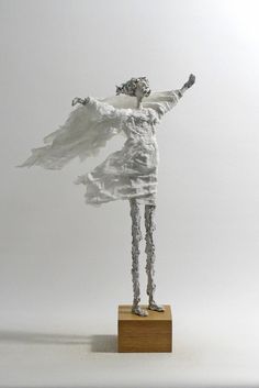 a white sculpture is standing on a wooden block and has its arms spread out in the air