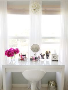 a white vanity with flowers and makeup on it in front of a window next to a sink