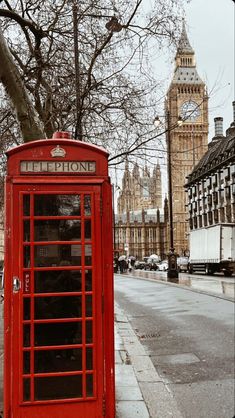 a red phone booth sitting on the side of a road next to a tree and clock tower