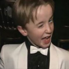 a young boy wearing a white suit and black bow tie in front of a chair