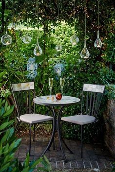 two chairs sitting at a table in the middle of a garden with hanging wine glasses