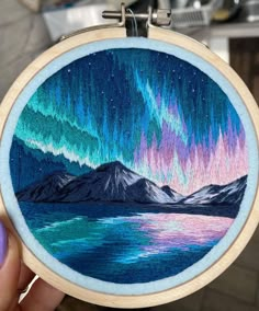 a person holding up an embroidery project with the aurora lights in the sky above mountains