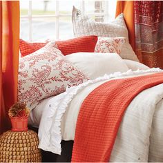 a bed with orange and white pillows in front of a window, next to a basket