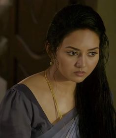 a woman with long black hair wearing a gray dress and gold necklaces, looking at the camera