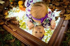 a baby looking at itself in a mirror with leaves on the ground and pumpkins behind it