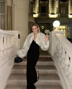 a woman in a black dress and white fur coat posing on the stairs at night