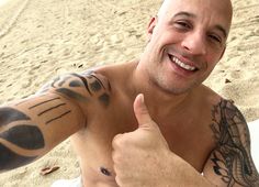 a bald man with tattoos giving the thumbs up sign on his arm at the beach