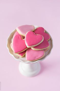 pink and white heart shaped cookies in a bowl on top of a pedestal against a pink background