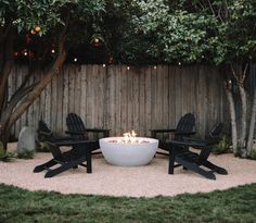 a fire pit surrounded by lawn chairs in a back yard with lights on the trees