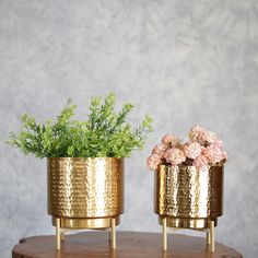 two gold planters with flowers in them sitting on a wooden table next to a wall