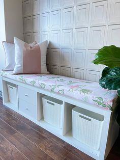 a white bench with three bins underneath it and a plant in the corner next to it