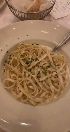 a white plate topped with pasta next to a bowl of bread and a glass of wine