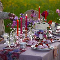 the table is set with flowers, candles and plates on it in front of an open field