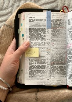 a person holding an open book with sticky notes attached to the pages, on top of a bed