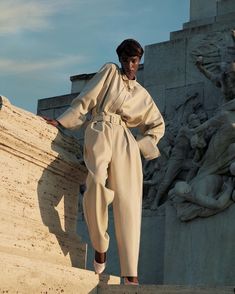 a woman standing on top of a stone wall next to a statue with her hands in her pockets