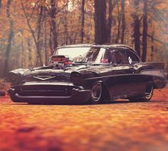 an old black car parked in the middle of a road with autumn leaves around it