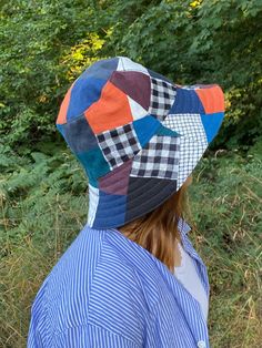 a woman wearing a patchwork hat standing in front of some bushes and trees with her back turned to the camera