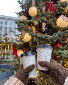 two people holding up coffee cups in front of a christmas tree with lights on it