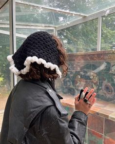 a woman in a black jacket is holding her hand up to the side of her head while wearing a crocheted hat