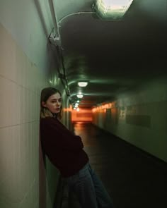 a woman leaning against a wall in a long hallway with light coming from the ceiling