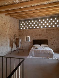 an empty bedroom with stone walls and exposed ceiling, bed in foreground is lit by light coming from the window