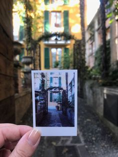 a person holding up a polaroid photo in front of an alleyway with green shutters