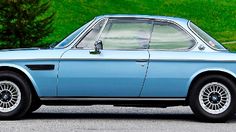 an old blue car is parked in front of some green grass and trees on the side of the road