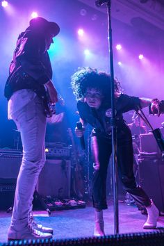 two people standing on stage with microphones in front of them and purple lights behind them