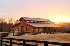 a barn with a fence in the foreground and trees in the background at sunset