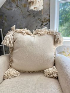 a white pillow with tassels on it sitting in a chair next to a window