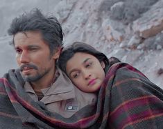 a man and woman wrapped in blankets standing next to each other on the side of a mountain