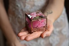 a person holding a small box with pink flowers in it's hands and wearing a wedding dress