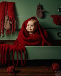 a baby wrapped in a red blanket sitting on top of a wooden bench next to balls of yarn