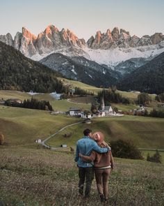 two people standing on top of a grass covered hillside with mountains in the back ground