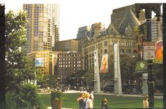 people are walking around in the city with tall buildings and green grass on both sides