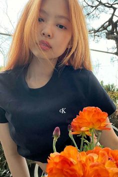 a woman with long red hair standing next to orange flowers and looking at the camera