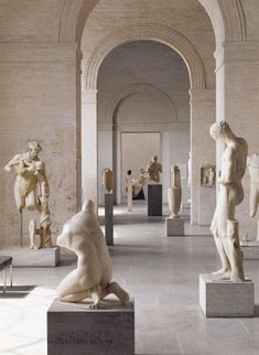 some statues are sitting on pedestals in a room with arches and columns, one is looking up at the ceiling