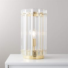 a clear and gold table lamp on top of a white counter with a gray wall in the background