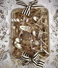 there are many forks and spoons in the tray on the lace tablecloth that is decorated with black and white ribbons