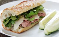 a sandwich with meat, lettuce and cheese on a white plate next to sliced cucumbers