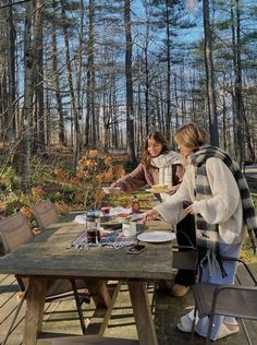 two women are sitting at a picnic table in the woods, one is serving food to the other