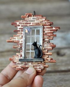 a hand holding a miniature cat in front of a brick wall with a small window