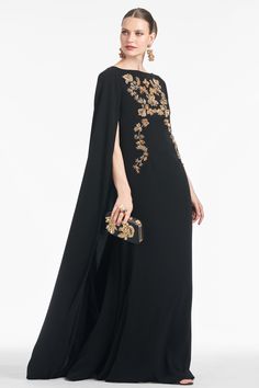 a woman in a black evening gown with gold embroidered cape over her shoulders and an embellished clutch bag