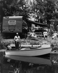 black and white photograph of man standing on boat next to trailer with rv in background