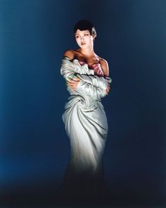 a woman in a white dress with her arms folded over her chest, standing against a blue background