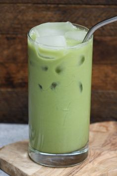 a glass filled with green liquid on top of a wooden board next to a spoon