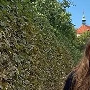 a woman walking down a street next to a tall hedge covered wall with a clock tower in the background