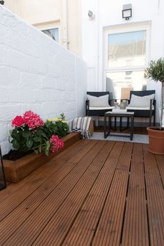 a wooden deck with potted flowers and chairs on the other side, next to a white brick wall