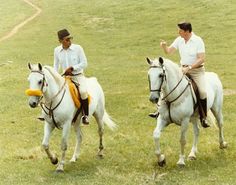 two men riding on the backs of white horses in a grassy field with one pointing