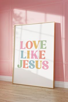 a white framed poster with the words love like jesus on it against a pink wall